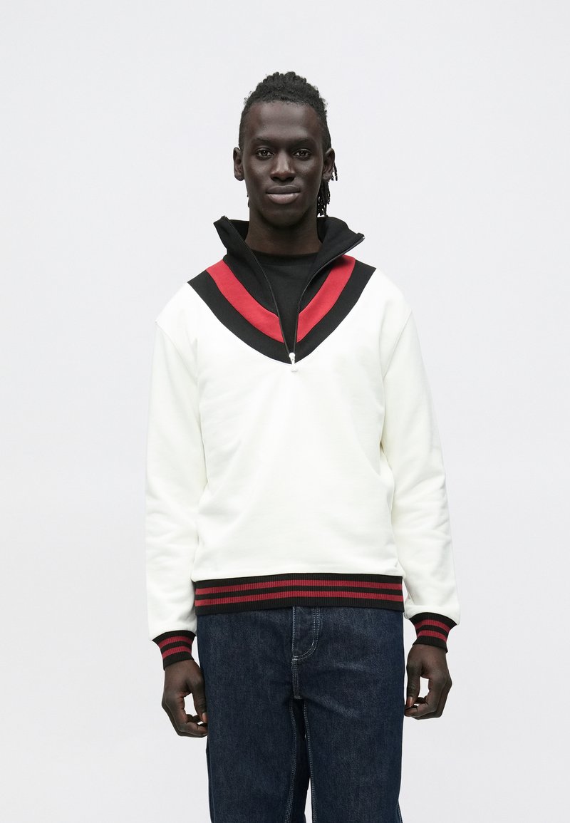 Valge lukuga dressipluus musta ja punase V-mustriga, soonikkoes mansettide ja äärega, koos tumelivate teksapükstega.