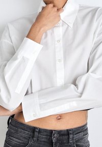 Camicia bianca a maniche lunghe con colletto, caratterizzata da una vestibilità regolare e una lunghezza leggermente accorciata. Dettagli con bottoni e polsini a contrasto.