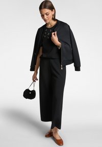 Giubbotto bomber nero con zip frontale, abbinato a un top nero con decorazioni, pantaloni larghi neri e mocassini marroni.