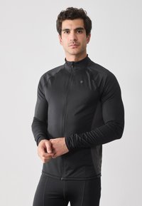 Chaqueta deportiva negra con cremallera, fabricada con un material elástico y suave. Diseño ajustado con mangas largas y cuello alto, cuenta con un pequeño logo en el pecho.