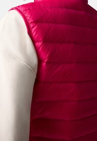 Fuchsiová nadýchaná vesta s horizontálním švy, vyznačující se hladkým povrchem a vysokým límcem, doplněná bílým dlouhým rukávem pod ní.