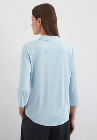 Light blue blouse with a collar, three-quarter sleeves, and a curved hemline. Smooth fabric with a subtle sheen. Back view only.