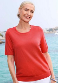 Koraalkleurig shirt met korte mouwen van gebreid materiaal, met een ronde halslijn en een rechte snit. Soepele textuur en geribbelde zoomdetails. Gedirigeerd met witte broeken.