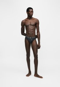 Homme athlétique à la peau foncée, debout pieds nus, portant un slip noir à motifs avec une ceinture à motif clé grecque, regardant vers sa gauche sur un fond uni.