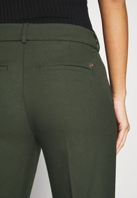 Gros plan d'une personne portant un pantalon vert olive sur mesure avec poches passepoilées à l'arrière et un haut noir côtelé.