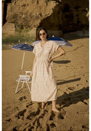 Blühendes Midikleid in Creme aus weichem Stoff, mit gerafftem Bund, kurzen Ärmeln und Knöpfen an der Vorderseite. Sandstrand mit gestreiften Sonnenschirmen.