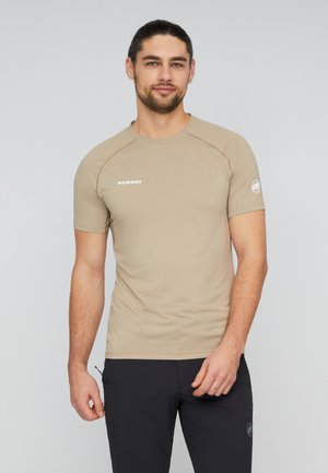 Mann mit dunklen Haaren und Bart, der ein beiges kurzärmeliges Mammut-Shirt und schwarze Hose trägt und vor einem schlichten hellen Hintergrund steht.