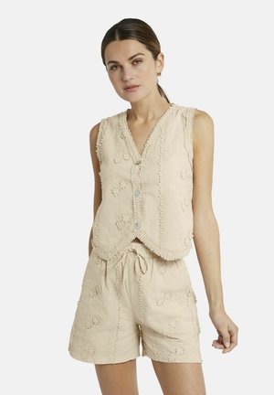 Frau in beigem ärmellosem, knöpfbarem Oberteil und passenden Shorts mit floral strukturierten Mustern, stehend vor einem schlichten weißen Hintergrund.