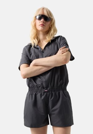 Blonde woman wearing a black short-sleeve romper with elastic waist and black wraparound sunglasses, standing with arms crossed.