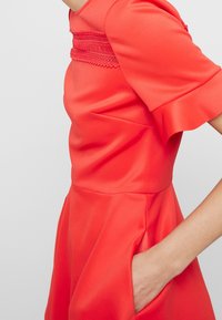 Robe en satin rouge avec des manches courtes, dotée d'un corsage texturé et de poches latérales. Texture lisse et taille cintrée menant à une jupe évasée.