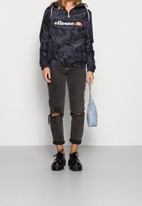 Mörk camouflagemönstrad vindjacka med framficka och logotyp, tillsammans med slitna, uppkavlade gråjeans och svarta plattformsskor. Håller en ljusblå väska.