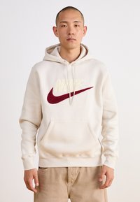 Beige hoodie tillverkad av mjukt tyg, med en stor lurvig vit "NIKE" logotyp och en vinröd swoosh. Inkluderar en framficka och justerbar huva med snörning.