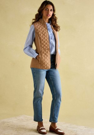 Beige gesteppte Weste mit hohem Kragen, kombiniert mit einem blauen Hemd, hellen Jeans und braunen Slippern mit weißen Streifen.