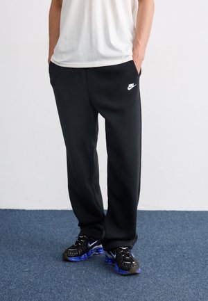 Mann trägt schwarze Nike-Sweatpants und schwarze Sneaker mit blauen Sohlen, steht auf blauem Teppich mit Händen in den Taschen.