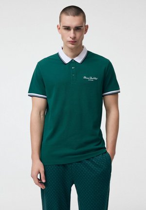 Młody mężczyzna z krótkimi włosami, ubrany w zieloną koszulkę polo z białym kołnierzykiem oraz ciemne zielone spodnie w wzory, stojący z jedną ręką w kieszeni.