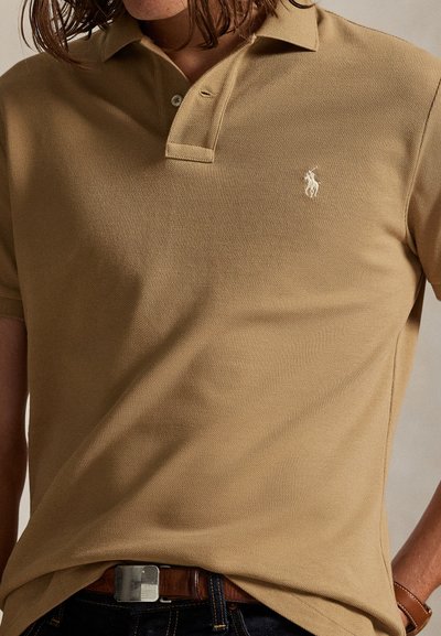 Moški nosi bež polo majico kratkih rokavov z majhnim belim logotipom konja, temne kavbojke, rjav pas s srebrno sponko in rjav ročni sat.