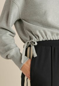 Svijetlosivi dukserica sa skupljenim strukom i vezicom, u kombinaciji s crnim trenirkama koje imaju bijele bočne pruge. Vidljiva meka tekstura tkanine.