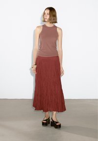 Top senza maniche marrone abbinato a una lunga gonna plissettata color ruggine. Sandali platform neri con suola spessa completano l'outfit. Tessuti lisci.