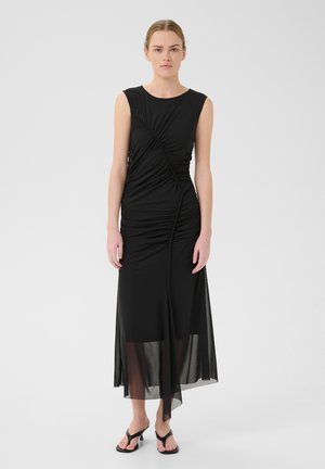 Vestito nero senza maniche con design arricciato, caratterizzato da un soprabito in rete trasparente, gonna spaccata e texture liscia. Indossato con semplici sandali neri.