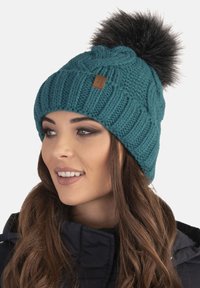 Gorro de punto en color verde azulado con un patrón de cable y un pompón de piel sintética negra. Presenta una etiqueta de cuero marrón en la parte delantera. Textura suave.