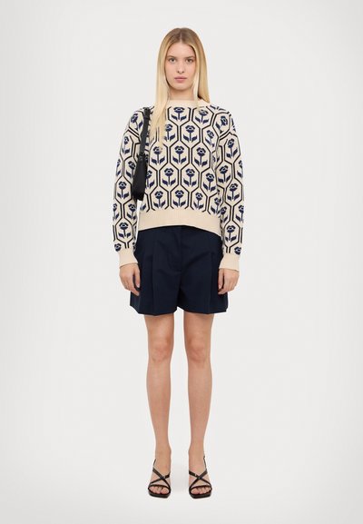 Pull en tricot à motif floral en bleu marine et crème, associé à un short plissé bleu marine et des sandales noires à lanières. Le modèle se tient devant un fond blanc.