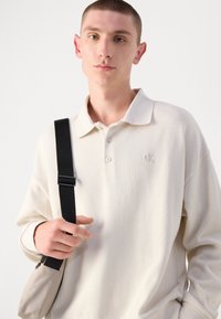 Helles beiges Strick-Polohemd mit Kragen, das zwei Knöpfe und ein dezentes Logo aufweist. Das Modell hält eine kleine Tasche mit einem schwarzen Riemen.