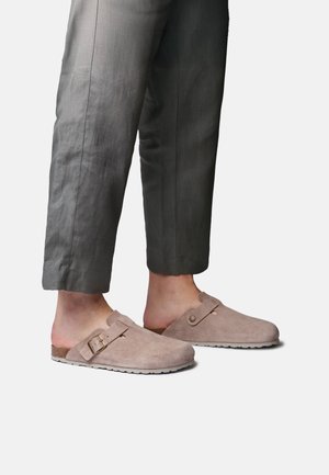 Cilvēks, kurš valkā pelēkas bikses un bēša krāsas zamšādas slip-on klogus ar regulējamām sprādzēm, stāv uz vienkārša balta fona.