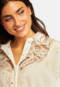 Blusa di lino off-white, con dettagli in pizzo intricati al colletto e davanti, chiusura con bottoni e accenti testurizzati.