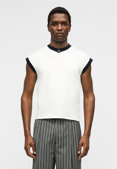 Άνδρας που φορά λευκή αμάνικη μπλούζα με μαύρη διακόσμηση και μαύρο-άσπρο κάθετα ριγέ παντελόνι, στέκεται μπροστά σε μονόχρωμο φόντο.