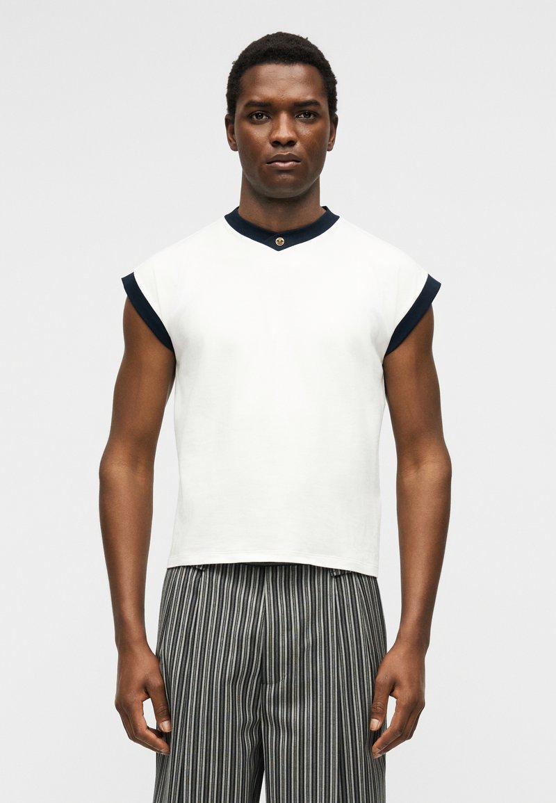 Man wearing white sleeveless top with black trim and vertically striped black and white pants, standing against a plain background.