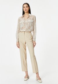 Lichte, doorschijnende beige blouse met knoopsluiting en twee borstzakken, gecombineerd met taillering crèmekleurige broek en crèmekleurige blokhak schoenen.