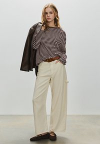 Ung kvinde iført brunstripet top, cremefarvede bukser med vide ben, mørkebrune sko, og hun holder en mørkebrun læderjakke over skulderen.