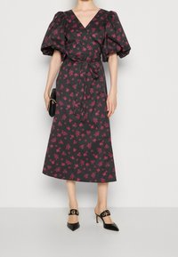 Vestido negro cruzado con estampado floral rosa, mangas abullonadas, escote en V y cintura con cinturón; combinado con tacones negros de punta afilada y un bolso de mano negro.