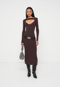 Smeđa pletena haljina rebrastog uzorka s dubokim izrezom na dekolteu, dugim rukavima i uskim krojem, uparena s crnim remenom i crnim čizmama do koljena.