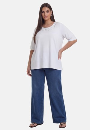 Kvinde iført hvid kortærmet top med dekorativ halsudskæring, vide blå jeans og sorte sandaler, stående med den ene hånd på hoften.