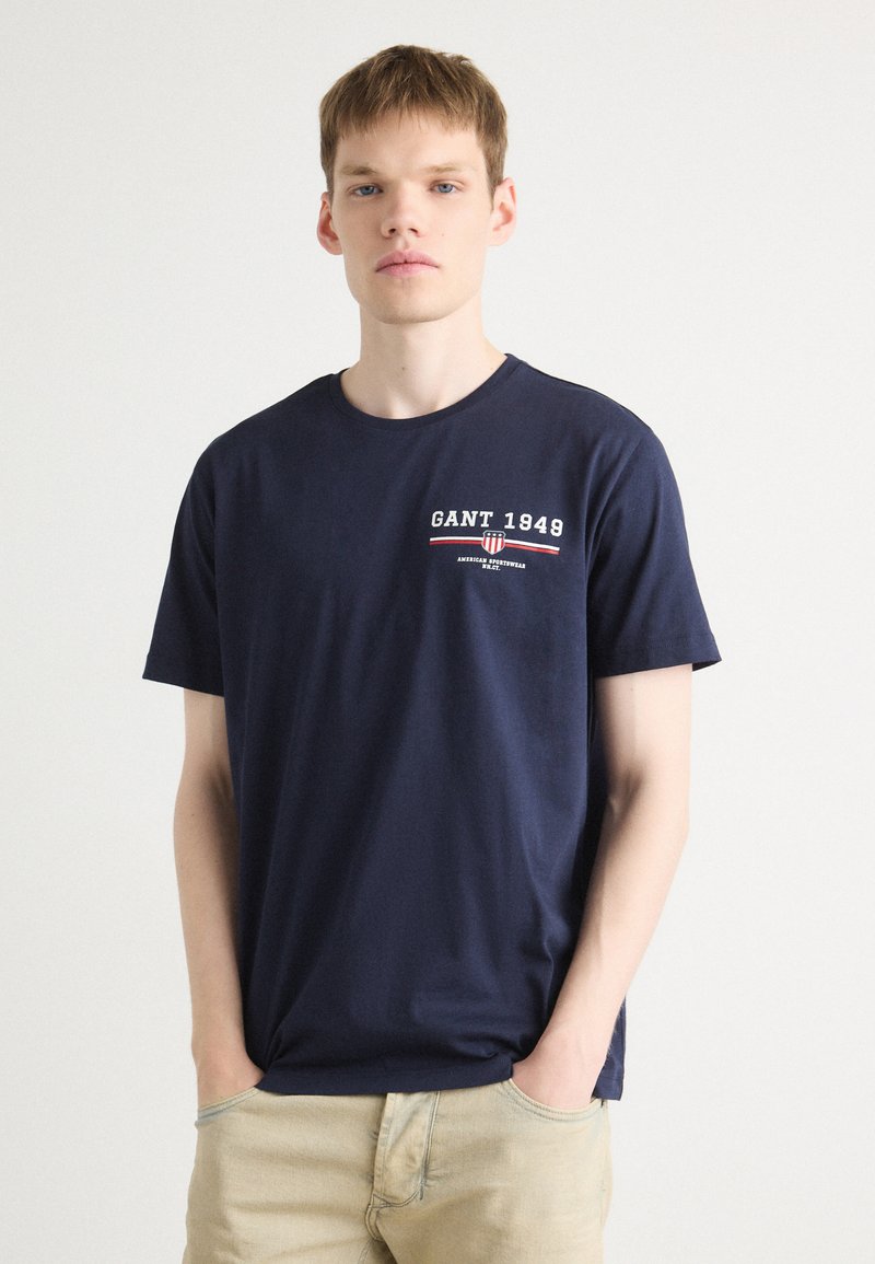 Marineblauwe katoenen T-shirt met "GANT 1948" geborduurd in wit en rood, klassiek ronde halslijn en korte mouwen.