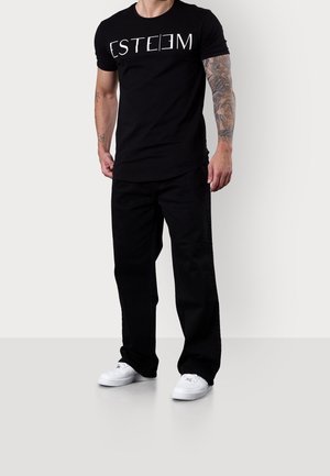 Hombre con camiseta negra con texto blanco "ESTEEM", pantalones negros, zapatillas blancas y tatuaje de manga completa en el brazo izquierdo, de pie sobre fondo liso.