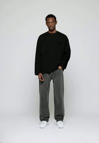 Uomo in camicia nera a maniche lunghe, jeans grigi larghi e sneakers bianche, in piedi contro uno sfondo grigio chiaro semplice.