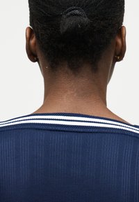 Femme à la peau foncée et aux cheveux tressés attachés en arrière, portant un haut côtelé bleu marine avec des doubles rayures blanches au niveau de l'encolure, vue de dos.