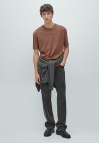 Bruin T-shirt met korte mouwen gecombineerd met antracietgrijze spijkerbroek; grijze trui om de taille geknoopt; zwarte schoenen, effen achtergrond.