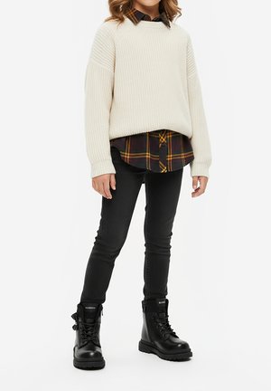 Dziecko ubrane w kremowy sweter z dzianiny na czarno-żółtej koszuli w kratę, czarne obcisłe jeansy i czarne sznurowane botki, stojące.