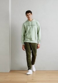 Felpa di cotone verde chiaro con la scritta "Copenhagen" stampata sul davanti, abbinata a pantaloni verdi oliva e sneakers bianche; in piedi su un pavimento di legno.