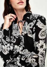Schwarze Bluse mit weißem Blumen- und Paisleymuster; verfügt über einen gerafften Ausschnitt mit einer Kordel und elastischen Bündchen. Leichter Stoff.