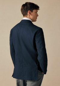 Marineblaues Sakko mit taillierter Passform, Schlitz hinten und Franselapel, kombiniert mit hellgrauen Hosen. Glatte Stofftextur.
