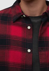 Camicia di flanella a quadri rossa e nera con bottoni bianchi, caratterizzata da un colletto con bottoni e un tessuto liscio e strutturato sotto.