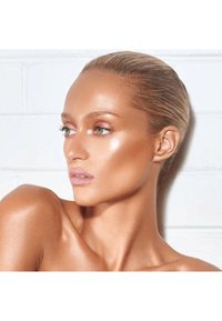 Retrato de una persona con piel bronceada, suave y radiante, cabello castaño claro y rasgos suaves, frente a un fondo de ladrillo blanco.