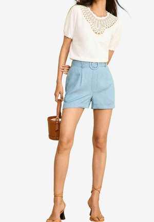 Kvinde iført hvid kortærmet top med blondedetaljer ved halsen, lyseblå bælteshorts, tanfarvede sandaler med stropper og med en brun spandtaske i hånden.