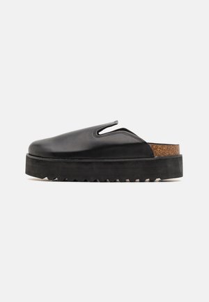 Chaussure slip-on en cuir noir avec bout rond, accent en liège et semelle en caoutchouc épaisse avec un bord dentelé. Détails découpés sur les côtés.
