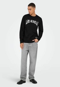 Felpa nera con testo bianco "LOOSE YOURSELF", scollo rotondo, maniche lunghe, polsini a costine, abbinata a jeans grigi a gamba larga e stivali neri.
