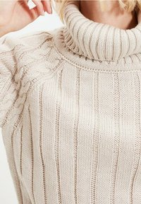 Close-up of a person wearing a cream-colored cable knit turtleneck sweater, showing textured vertical and braided knit patterns.